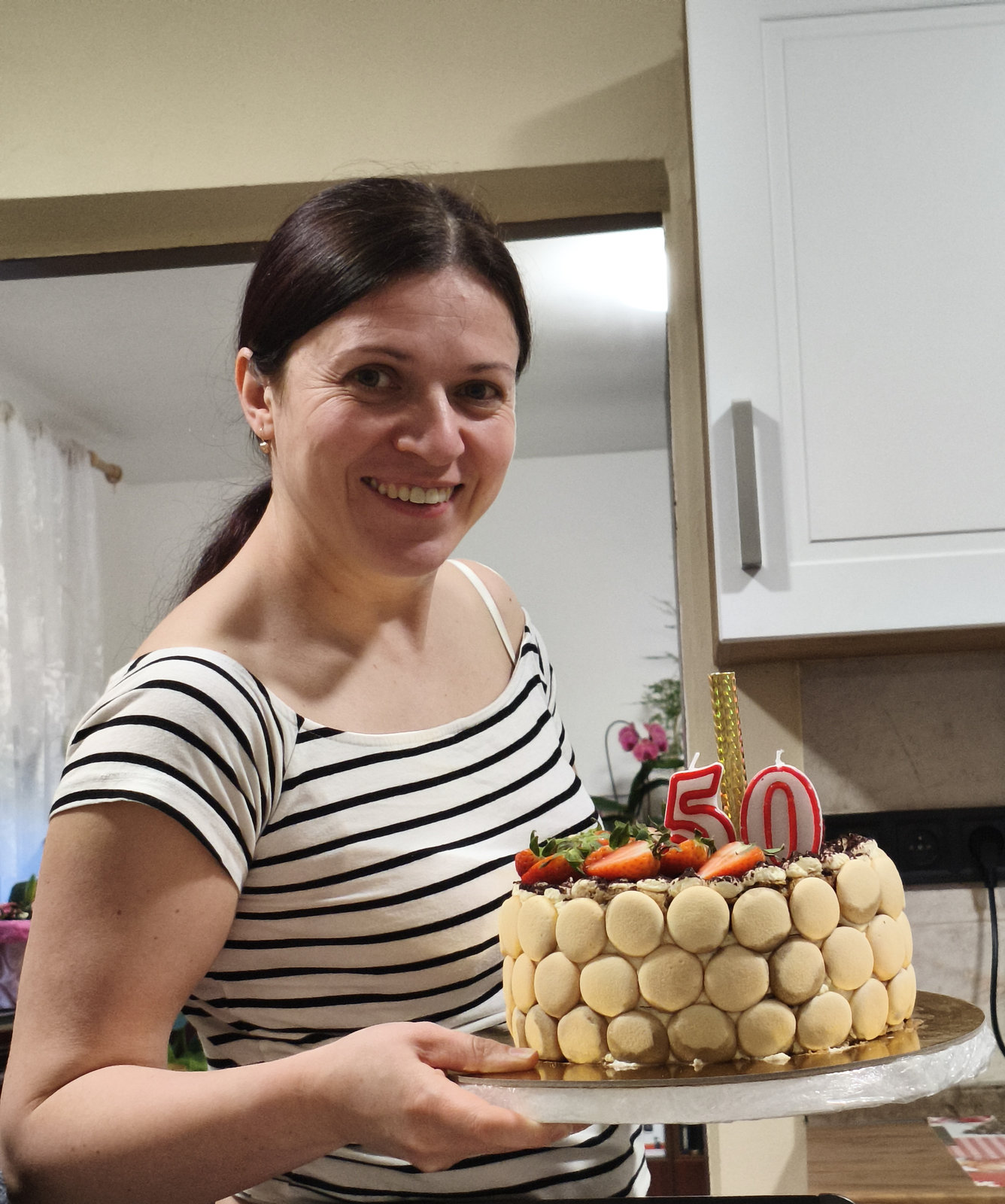 Usměvavá žena drží narozeninový dort s číslem 50, zdobený ovocem a makronkami.