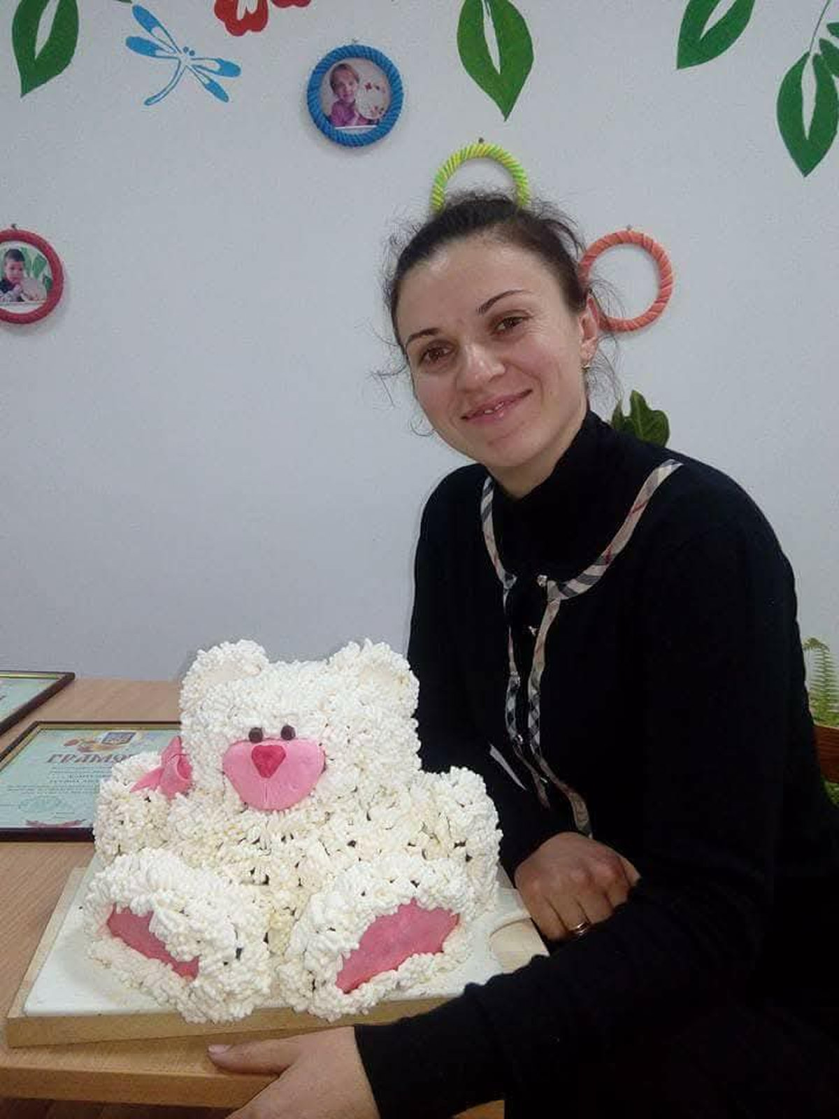 Žena sedí u stolu a drží velký dort ve tvaru medvídka s růžovou polevou.